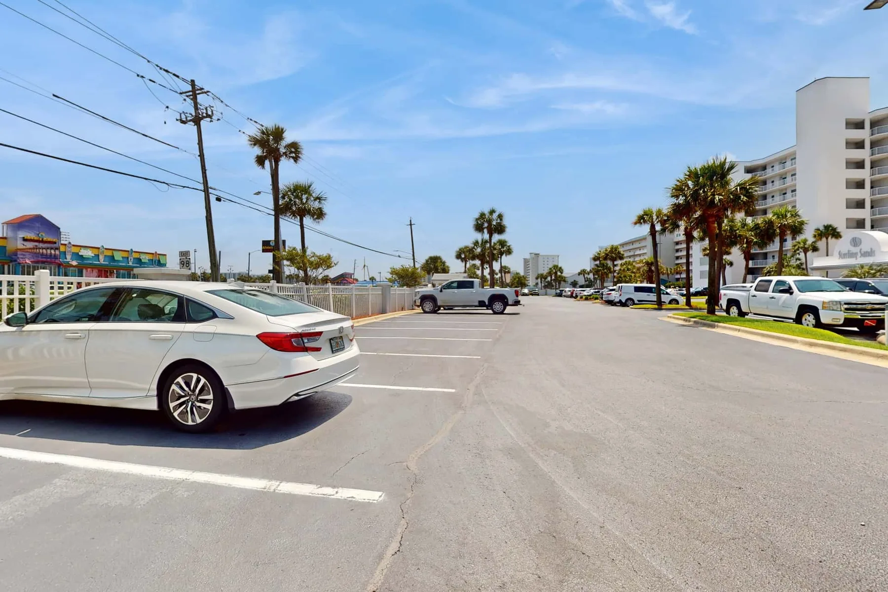 A paved parking lot with parked cars and palm trees beside a hotel.