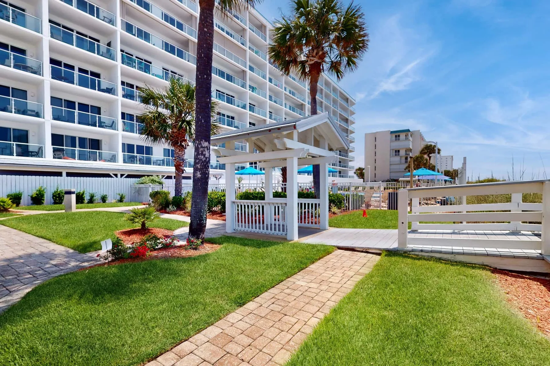 A landscaped outdoor walkway passes a white pergola beside a multi-story building.