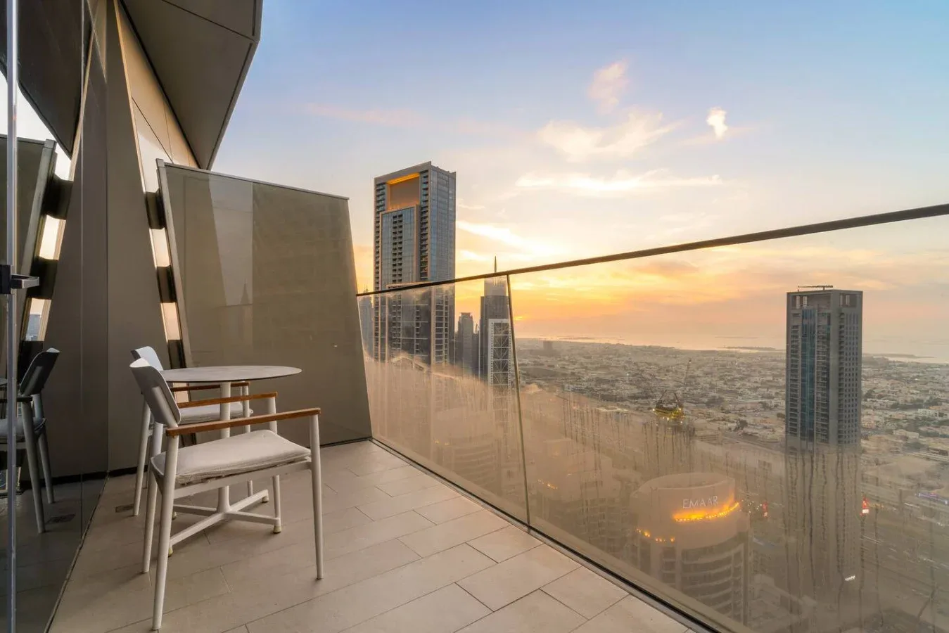 A balcony with seating overlooks nearby high-rise buildings at sunset.