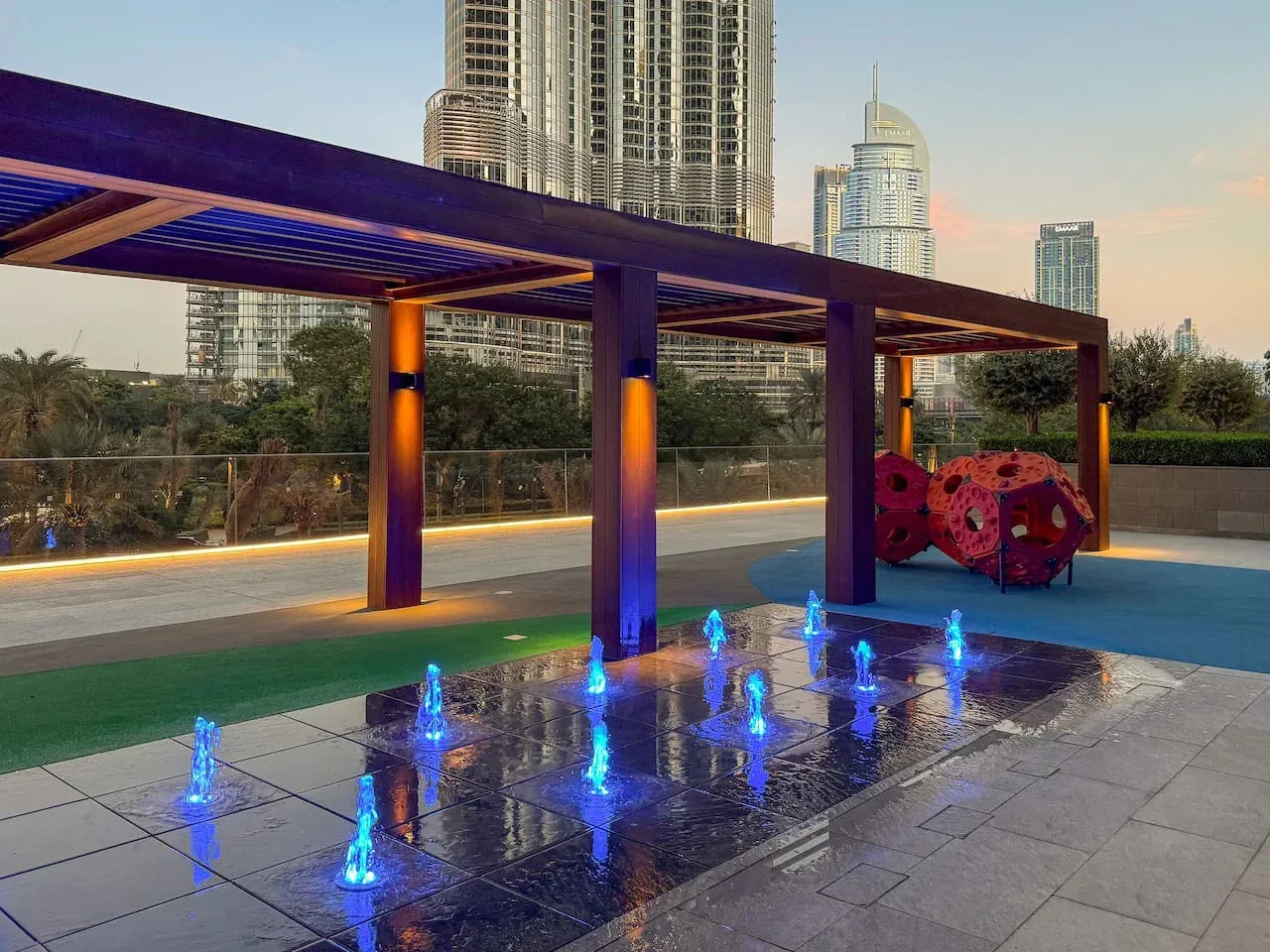 A rooftop terrace features a pergola and blue-lit fountains, with city buildings in the background.