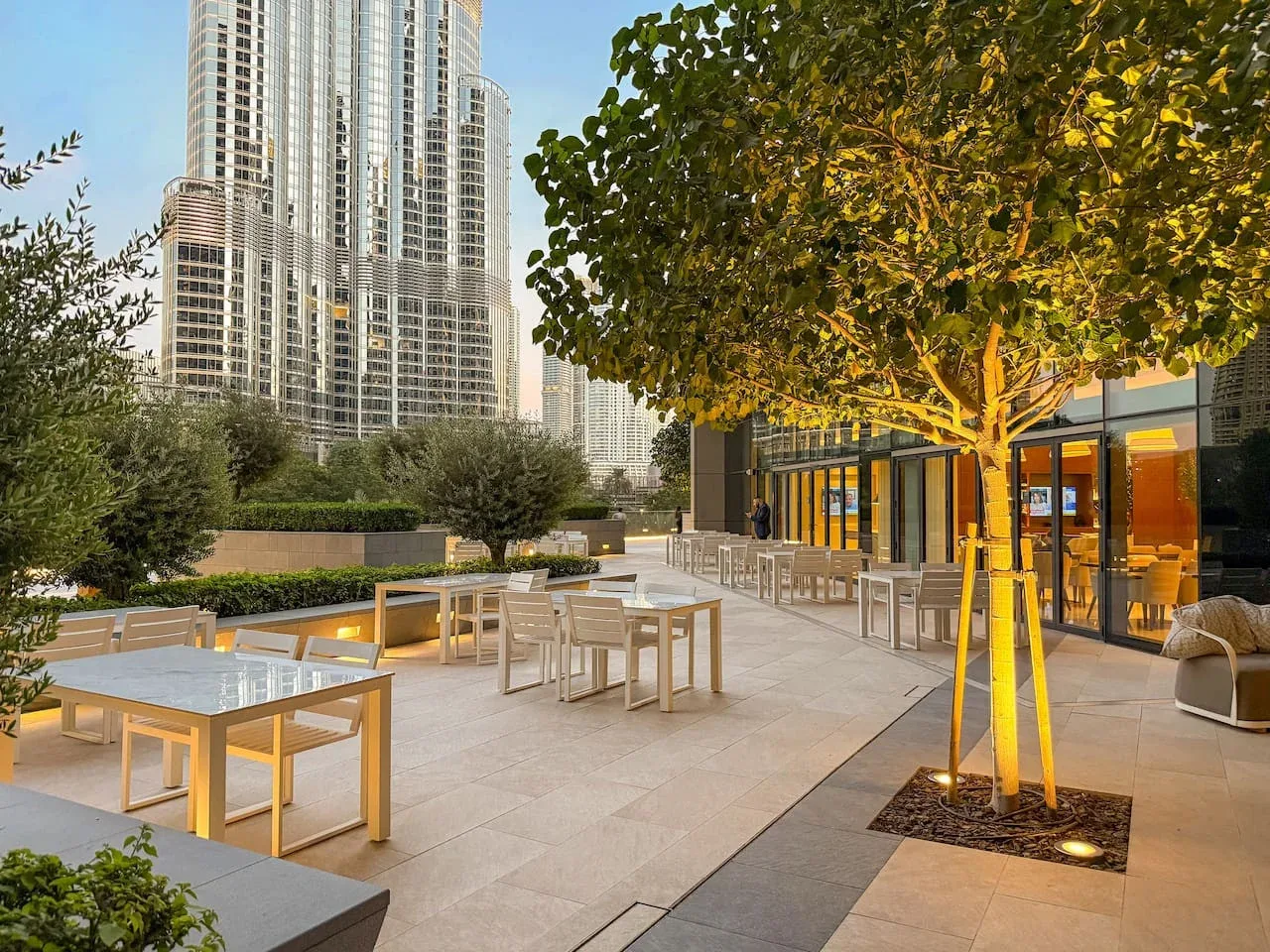 An outdoor terrace with dining tables, seating, and a tree beside a glass-fronted building.