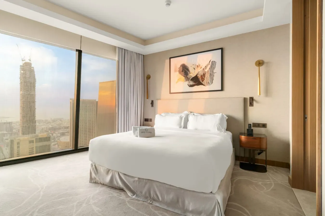Un dormitorio luminoso con una cama bien tendida junto a grandes ventanales con vistas a la ciudad.