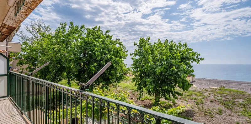 A balcony overlooks leafy trees and a sandy shoreline beside the sea.