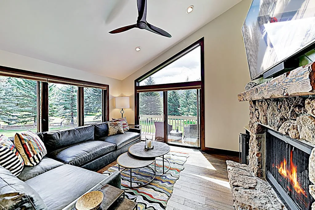 A bright living room with seating, large windows, and a stone fireplace.