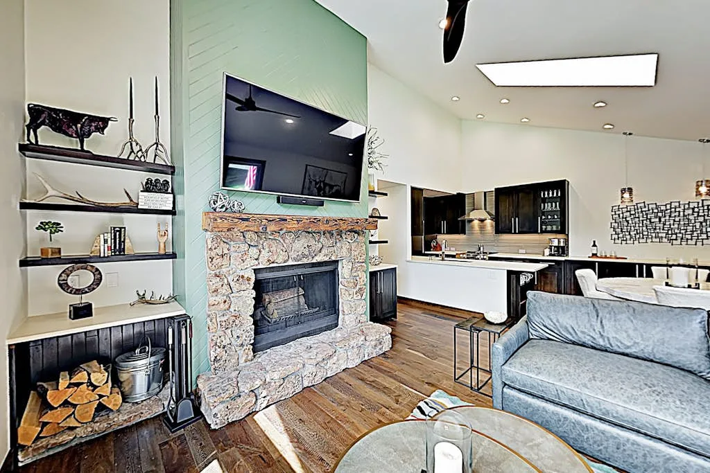 A living room with a stone fireplace, television, and seating beside an open kitchen.