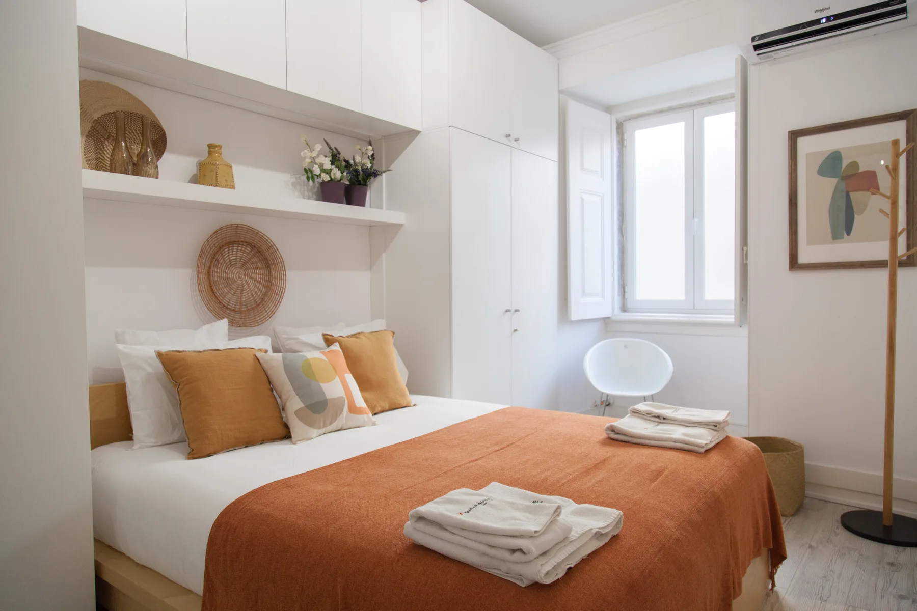 Un dormitorio luminoso con ropa de cama blanca, textiles color óxido y estantes empotrados.