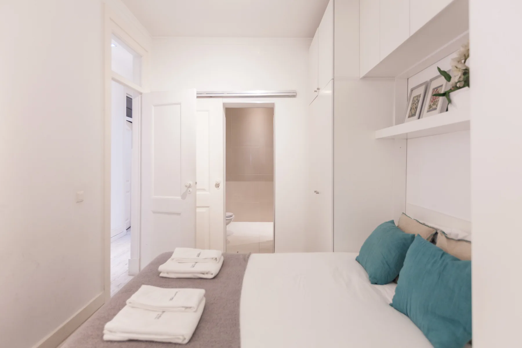 Dormitorio luminoso con una cama, toallas dobladas y estantes empotrados.