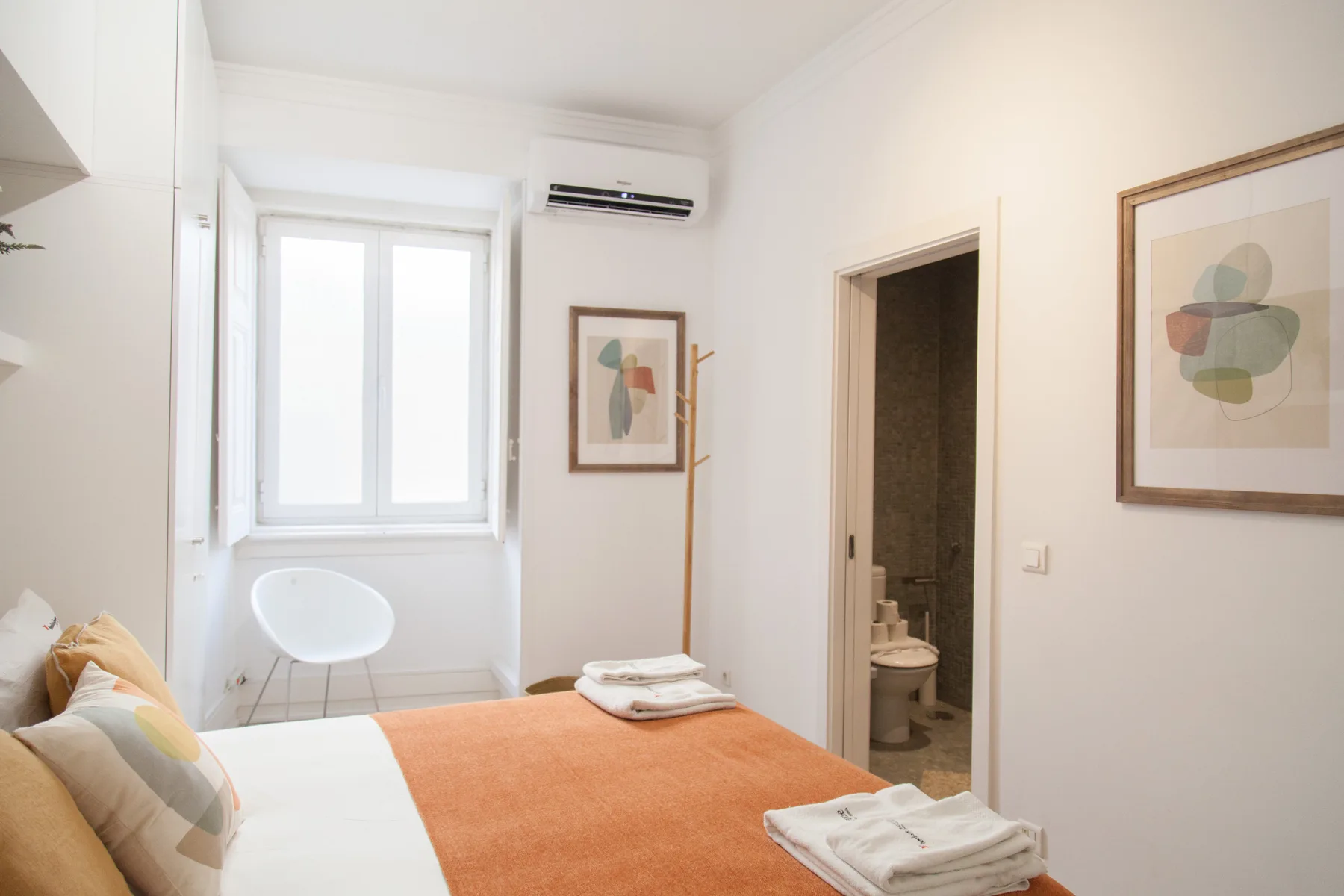 Un dormitorio luminoso con una cama, cuadros enmarcados y la puerta abierta al baño.