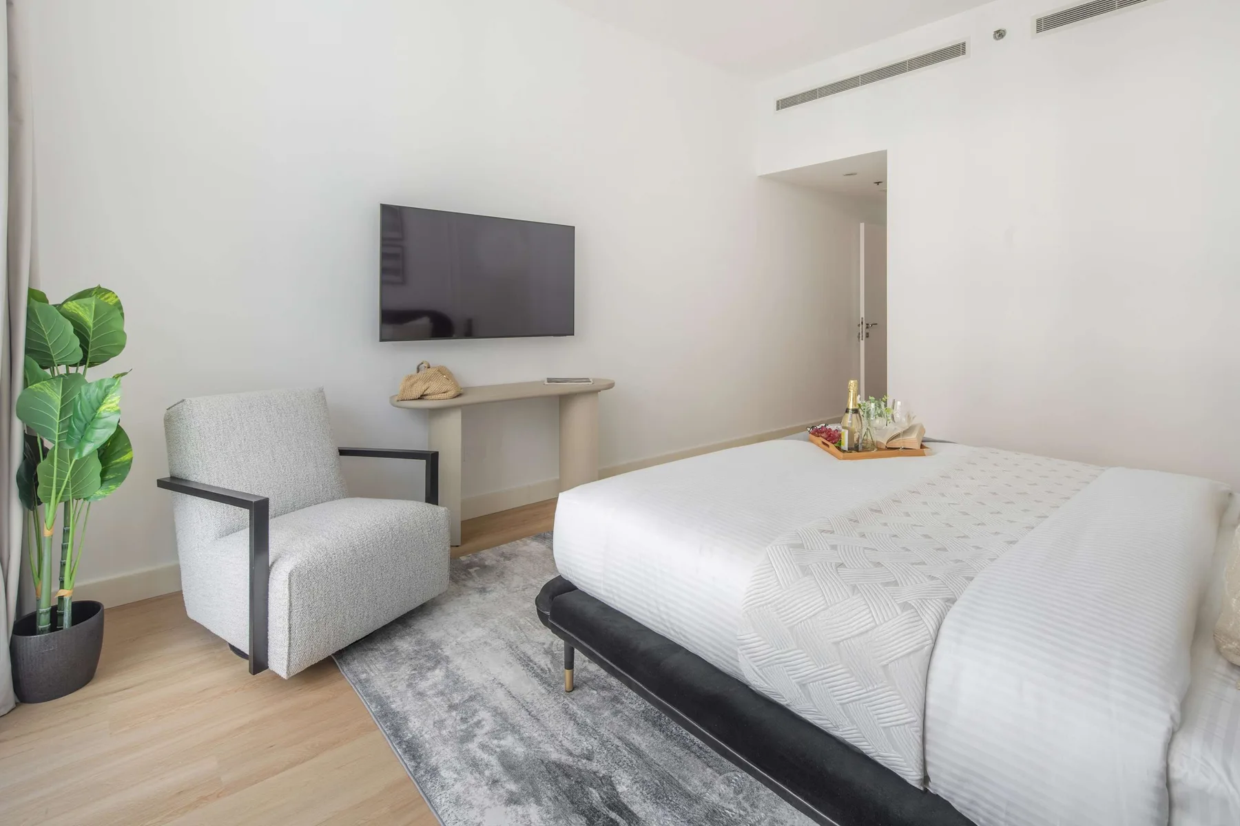 A bright bedroom with a neatly made bed, a chair, and a wall-mounted television.
