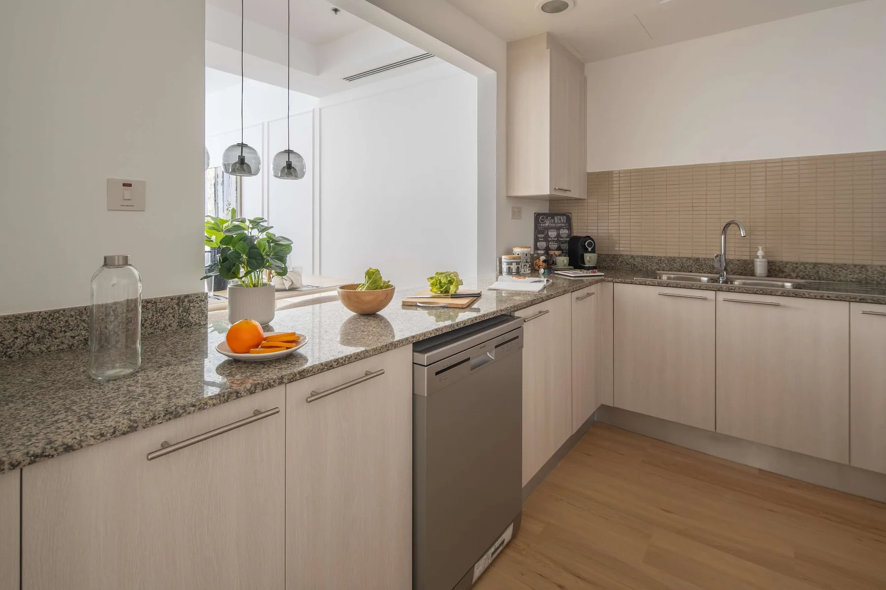 A bright kitchen with pale cabinets, a stone countertop, and a stainless steel dishwasher.