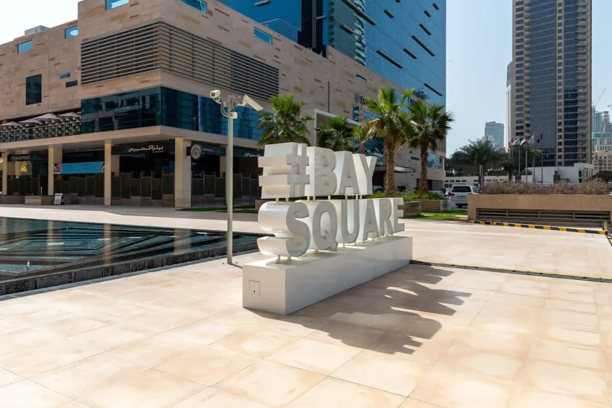A sunlit plaza features a large BAY SQUARE sign beside a reflecting pool.