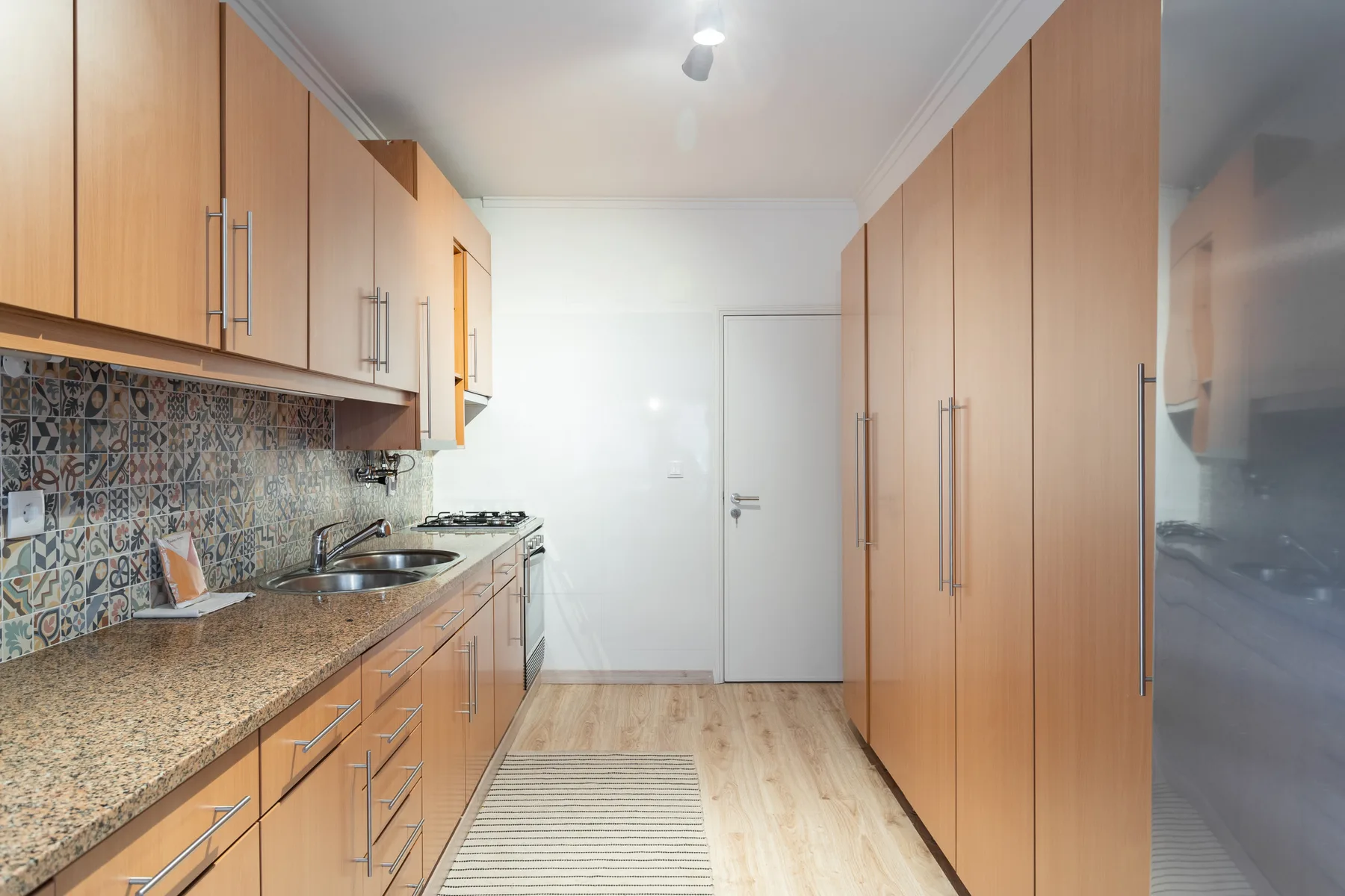 A narrow kitchen with wooden cabinets, a patterned backsplash, and a long counter.
