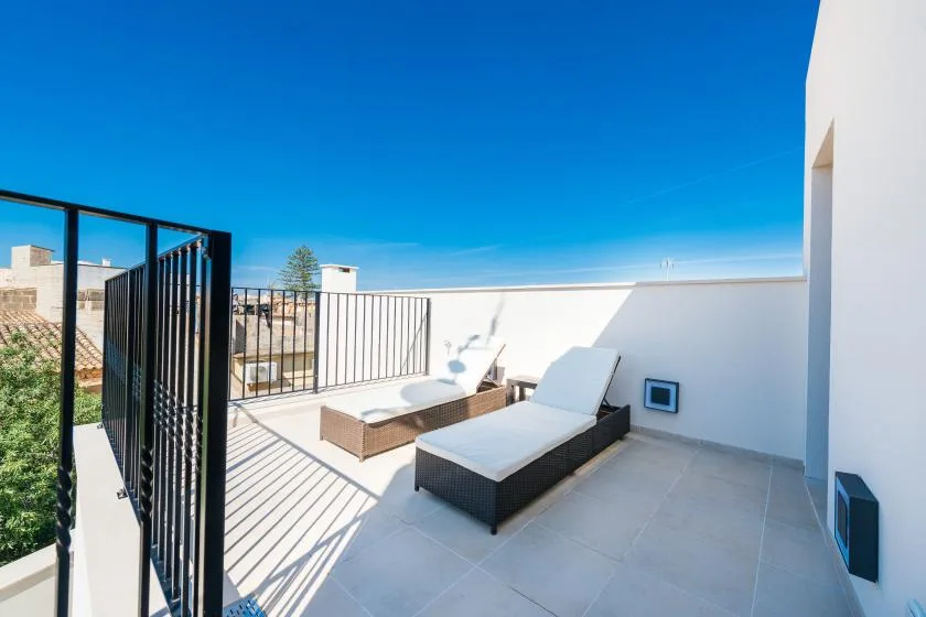 A sunny rooftop terrace has lounge seating, pale tiled flooring, and a metal railing.
