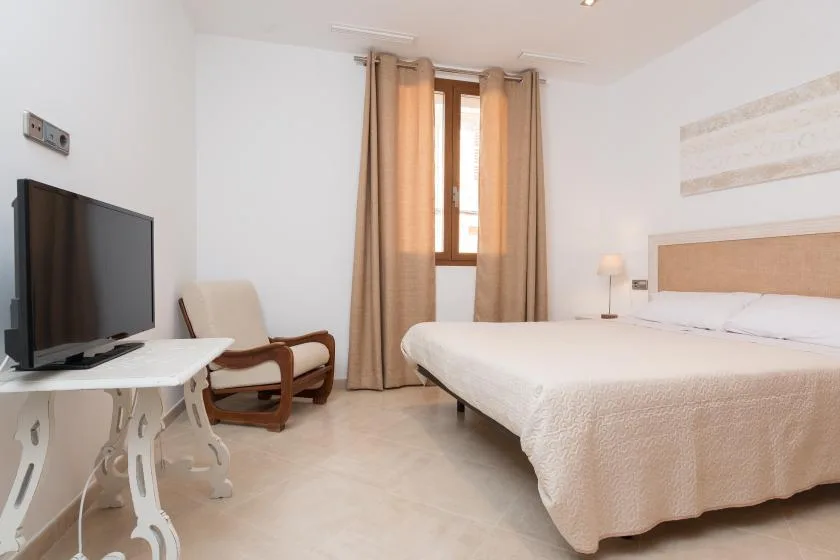 A bright bedroom with a large bed, a television, and a curtained window.