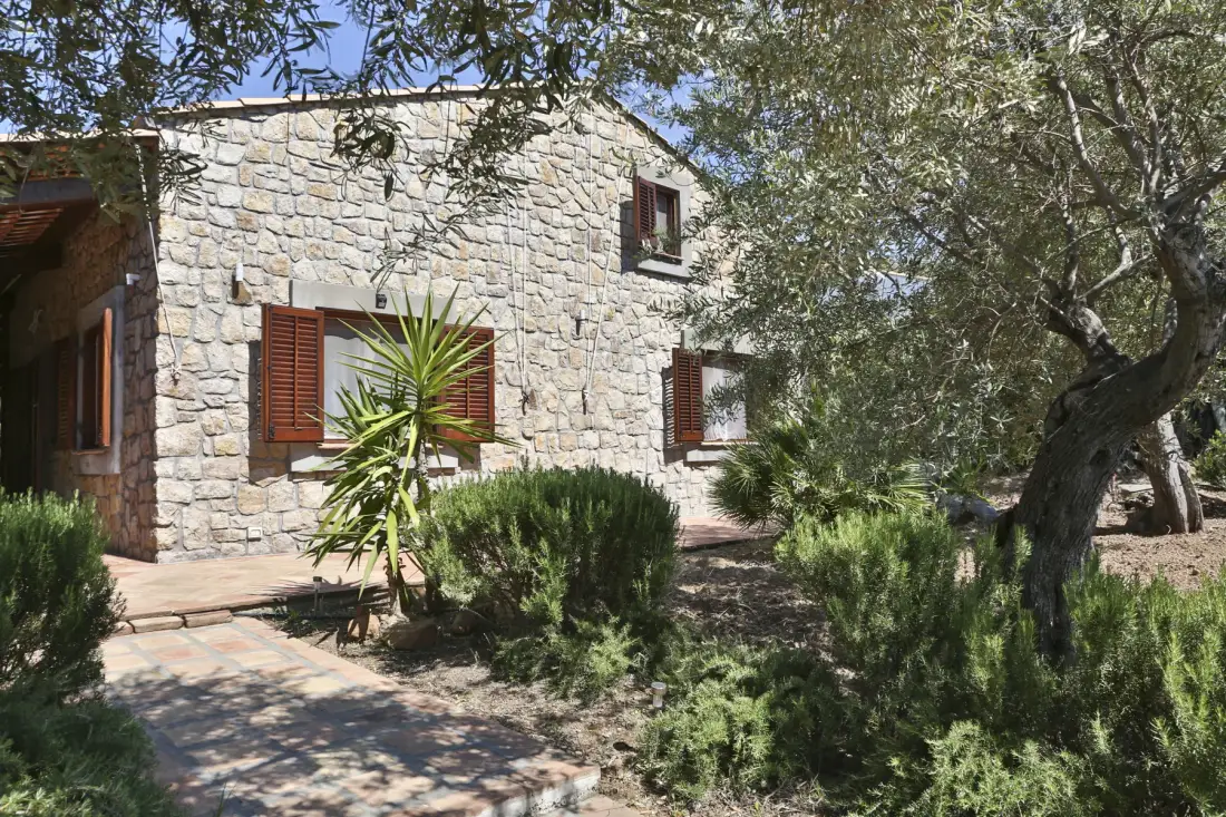 A stone house stands among trees and shrubs beside a paved walkway.