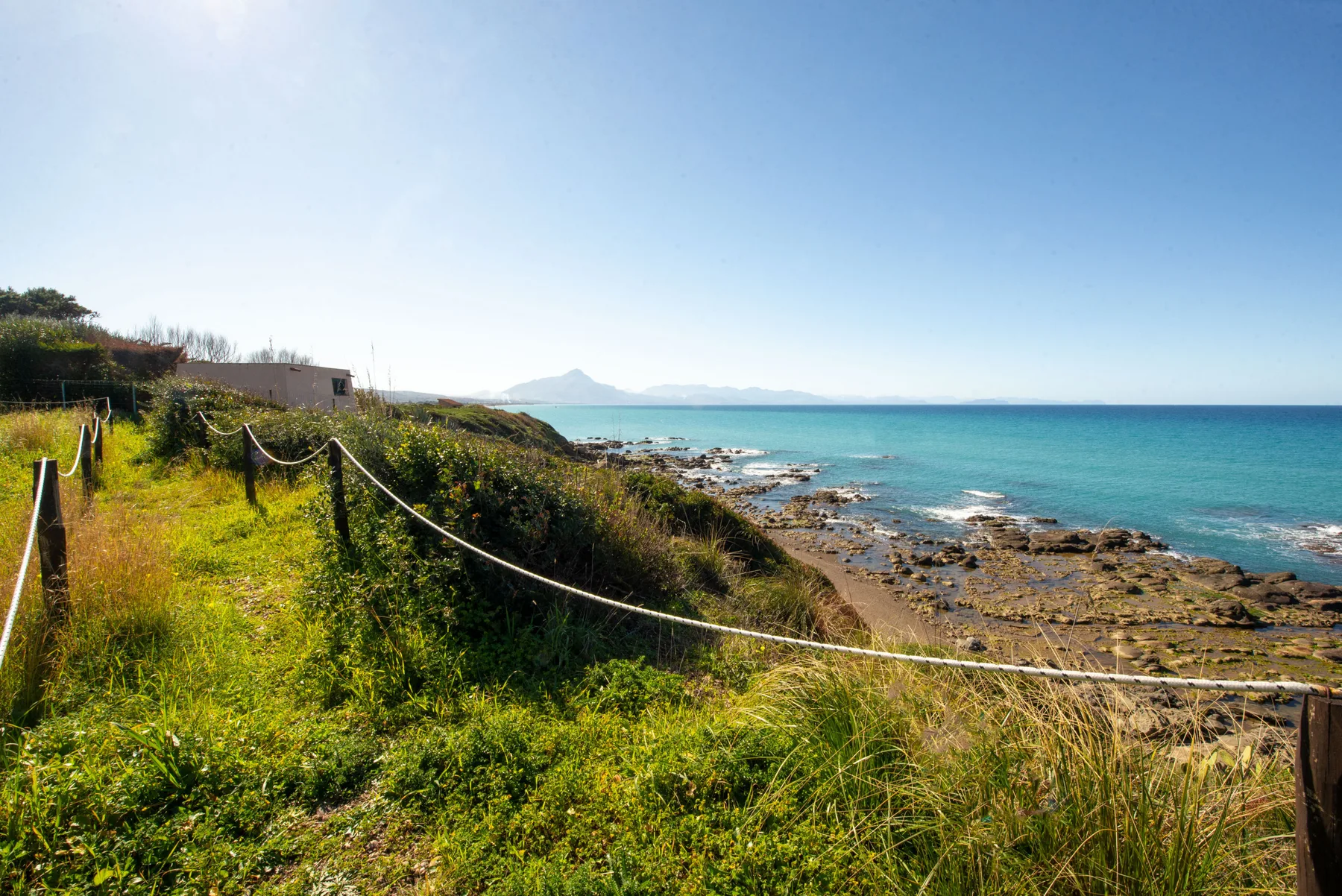 A grassy coastal path follows a rocky shore beside the blue sea.