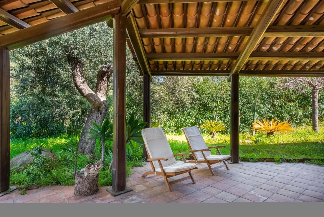 A covered patio with two reclining seats overlooks a green garden.