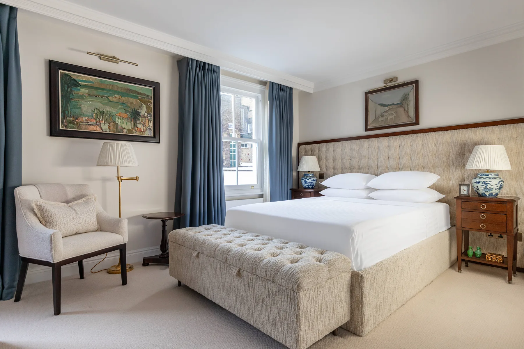 A bedroom with a large bed, upholstered bench, and seating beside a curtained window.