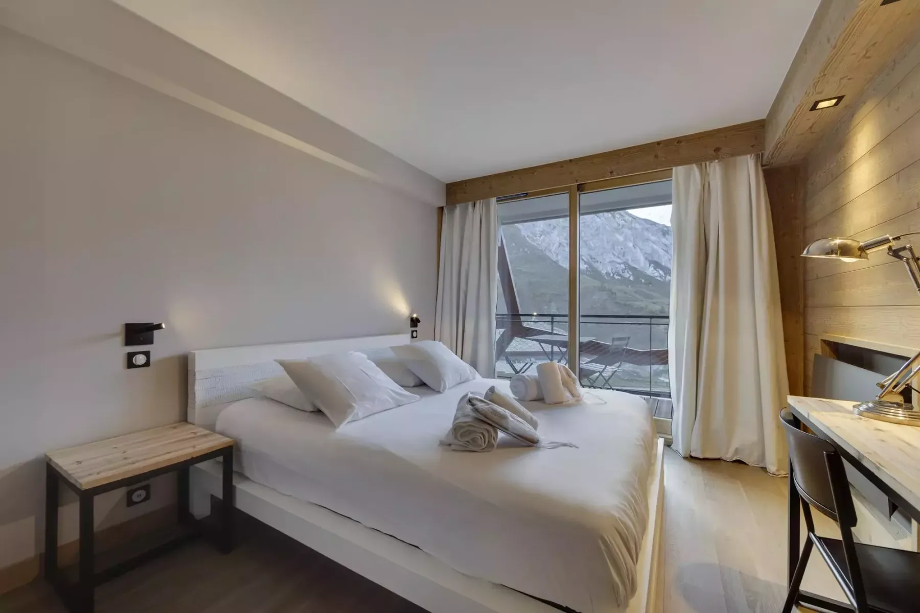 A bedroom with a bed beside glass doors opening onto a balcony and mountain view.