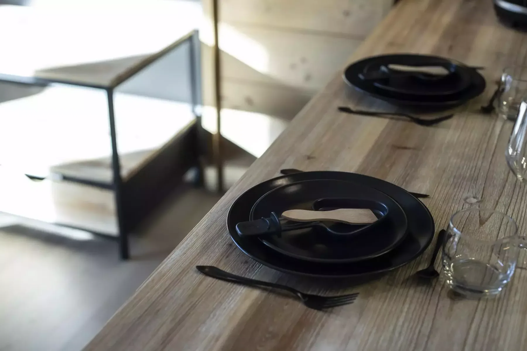 A wooden dining table is set with dark plates and glassware.