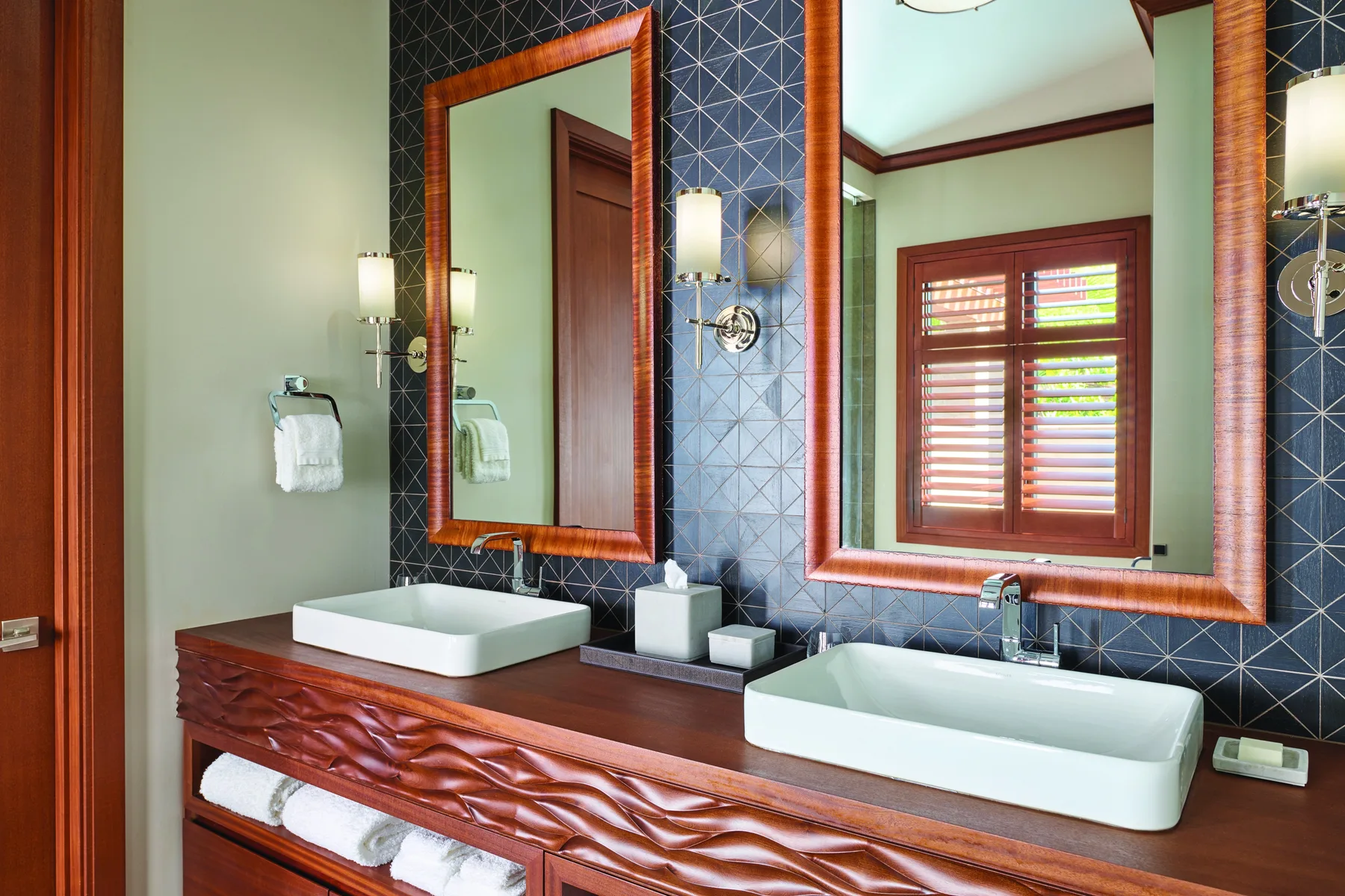 Un tocador de baño con dos lavabos, espejos enmarcados y una ventana con persianas de madera.