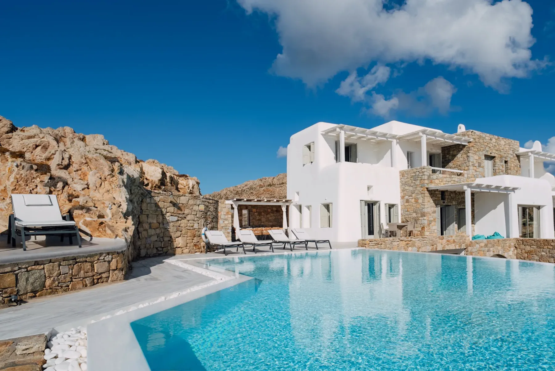 A swimming pool beside a white building and stone walls under a blue sky.