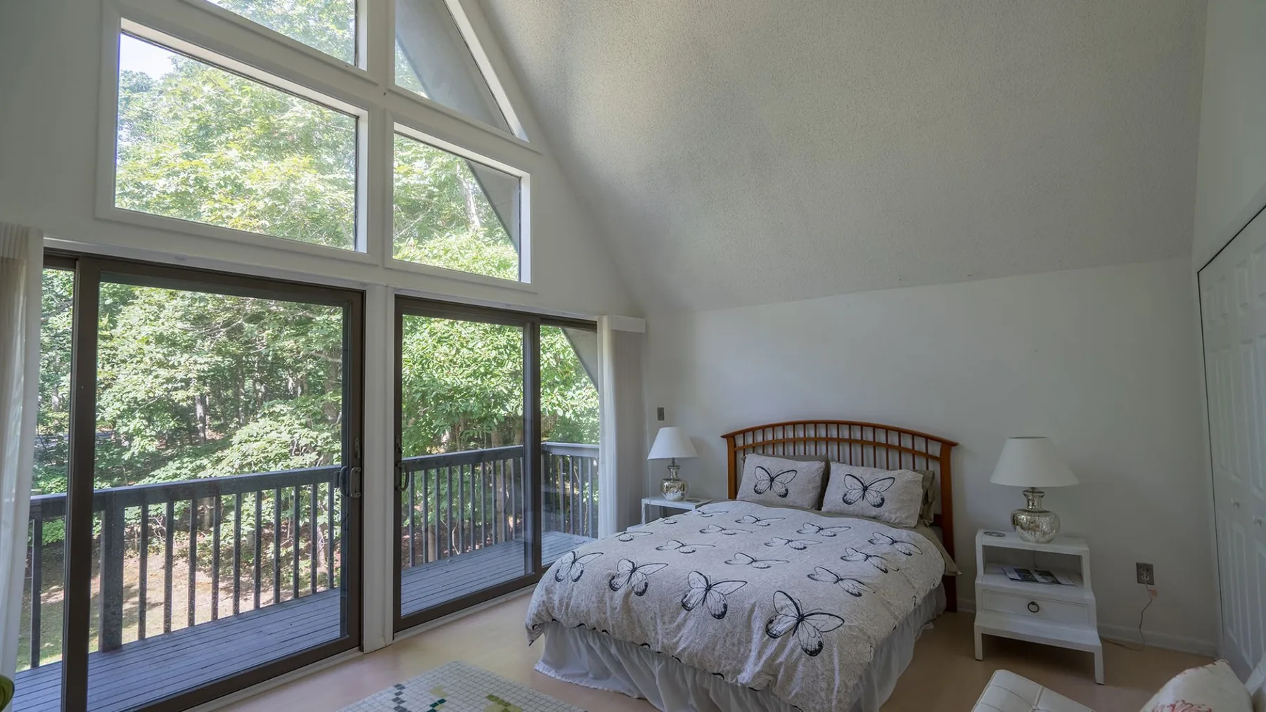 A bright bedroom with a bed beside large windows overlooking trees.