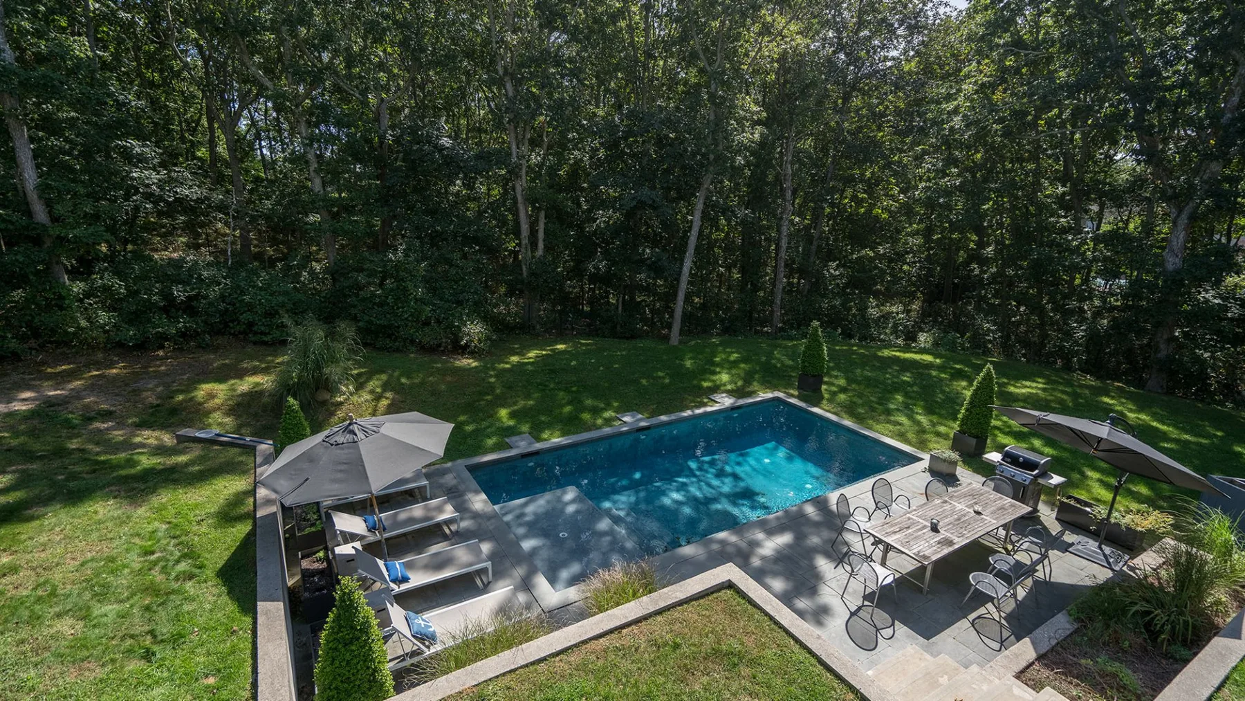 Una piscina al aire libre está junto a un patio pavimentado rodeado de árboles.