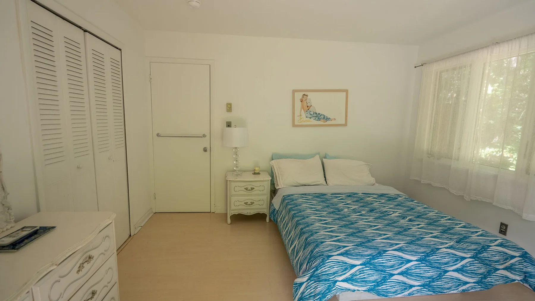 A bedroom with a bed covered in a blue patterned bedspread beside a window.
