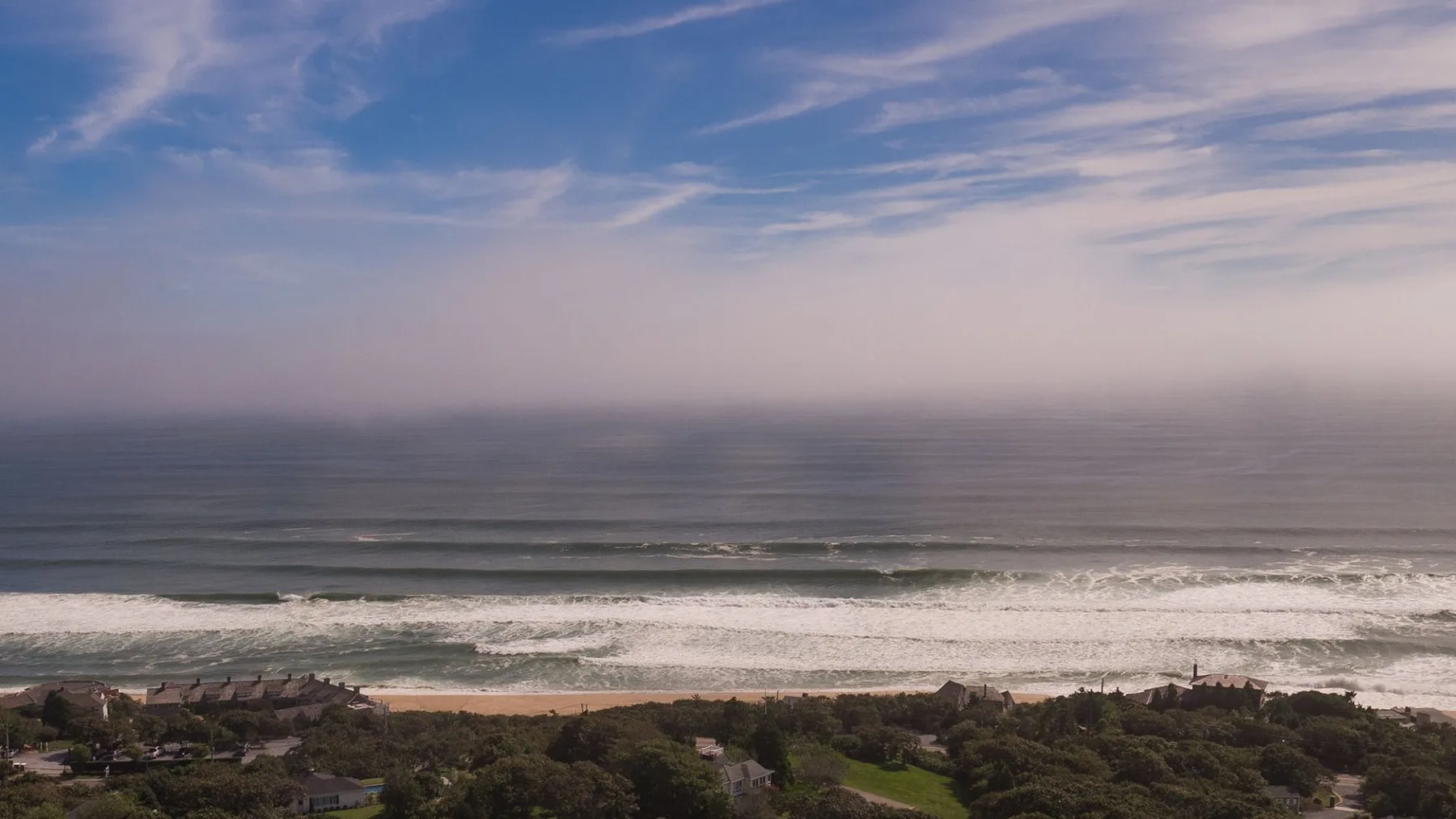 A coastal landscape with ocean waves, a sandy beach, and buildings among greenery.