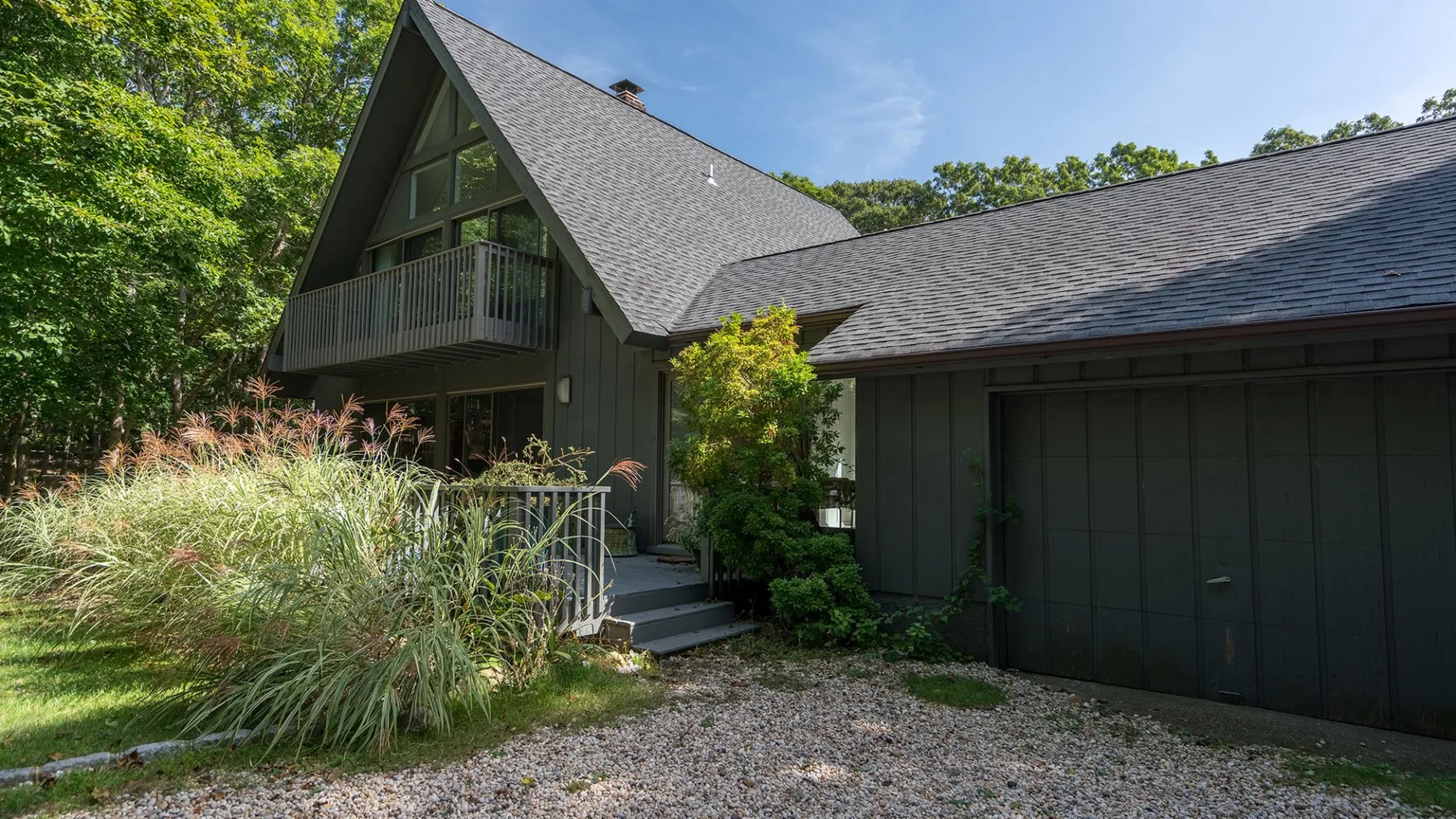 A dark-sided house with a steep gabled roof and a small balcony sits among trees.