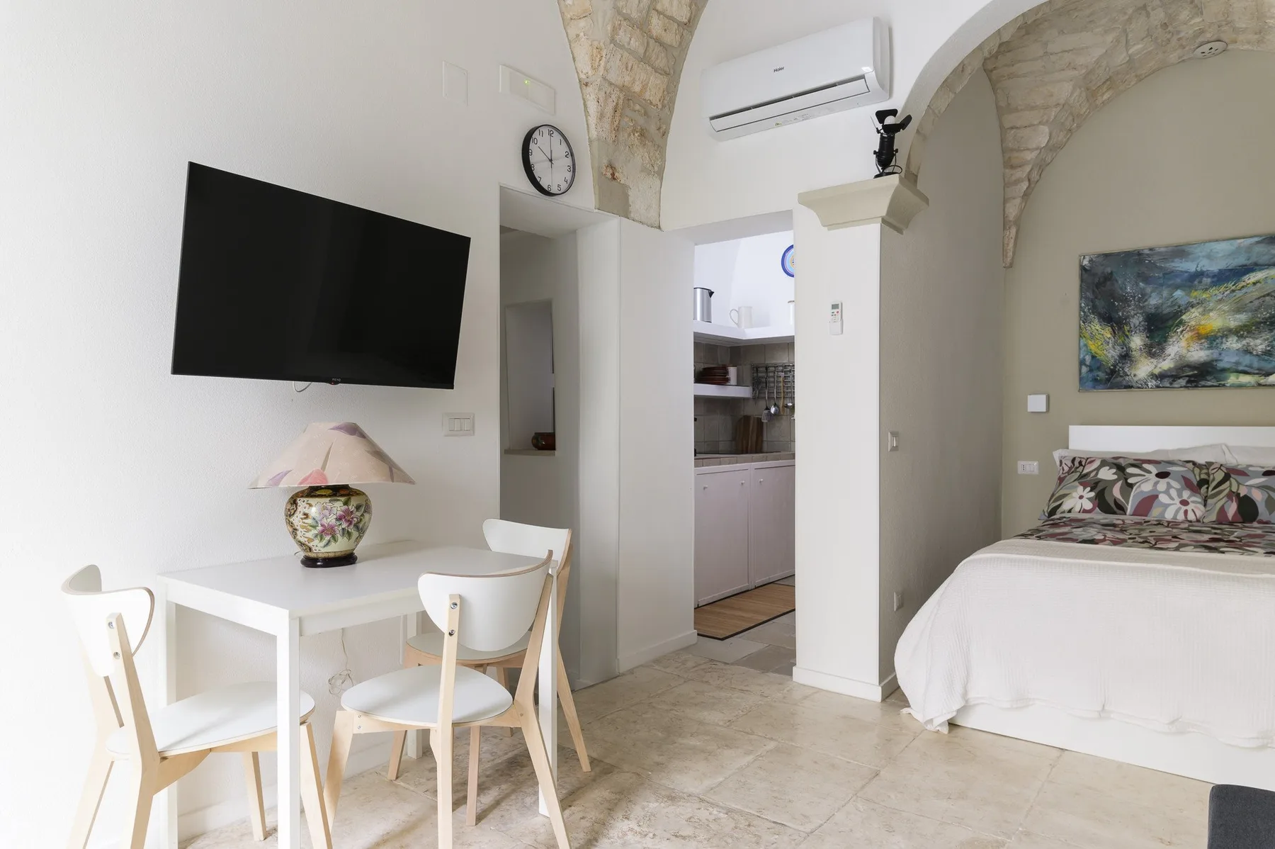 A bedroom with a dining table, wall-mounted television, and an open kitchen area.