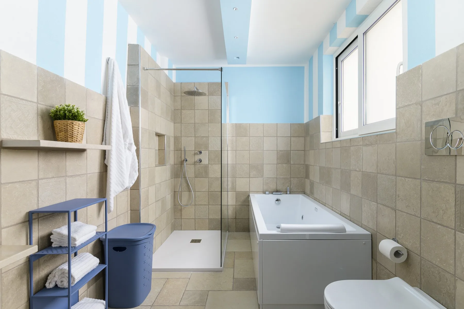 A bright tiled bathroom with a glass shower, bathtub, and large window.