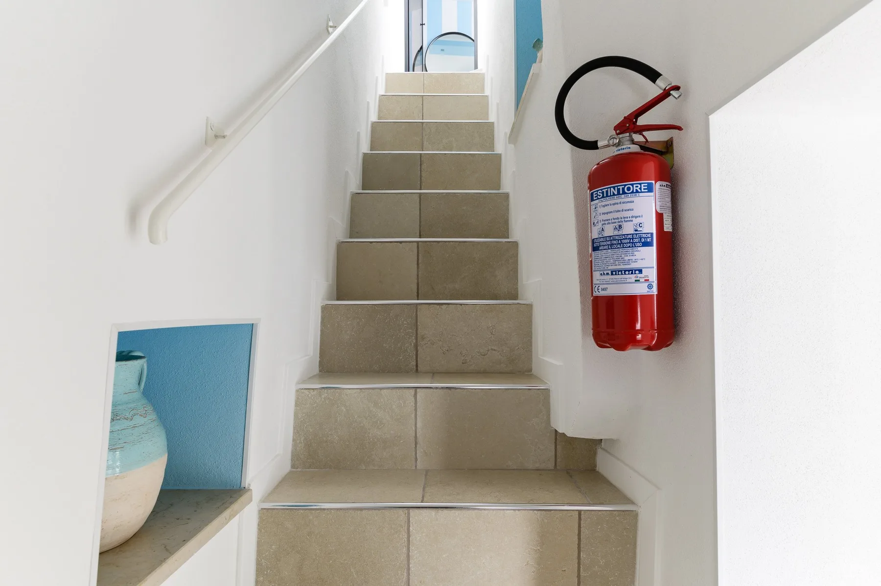 A tiled staircase rises between white walls, with a fire extinguisher mounted beside it.