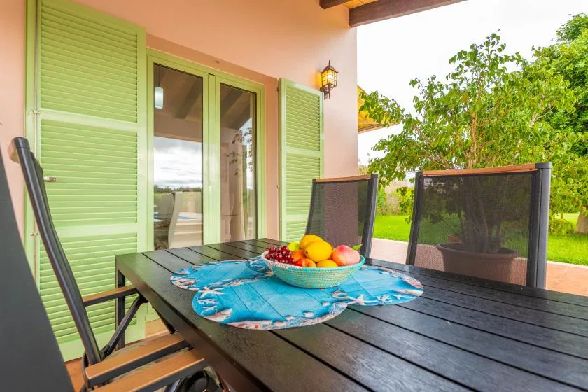 A covered outdoor dining area with a table set beside open green shutters.