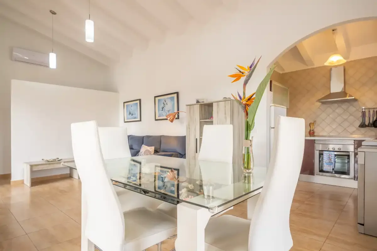 A bright dining area with glass table and white seating opens into a kitchen.