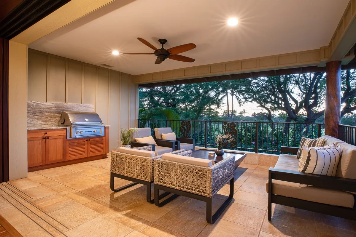 A covered patio has seating, a table, and a built-in grill overlooking trees.