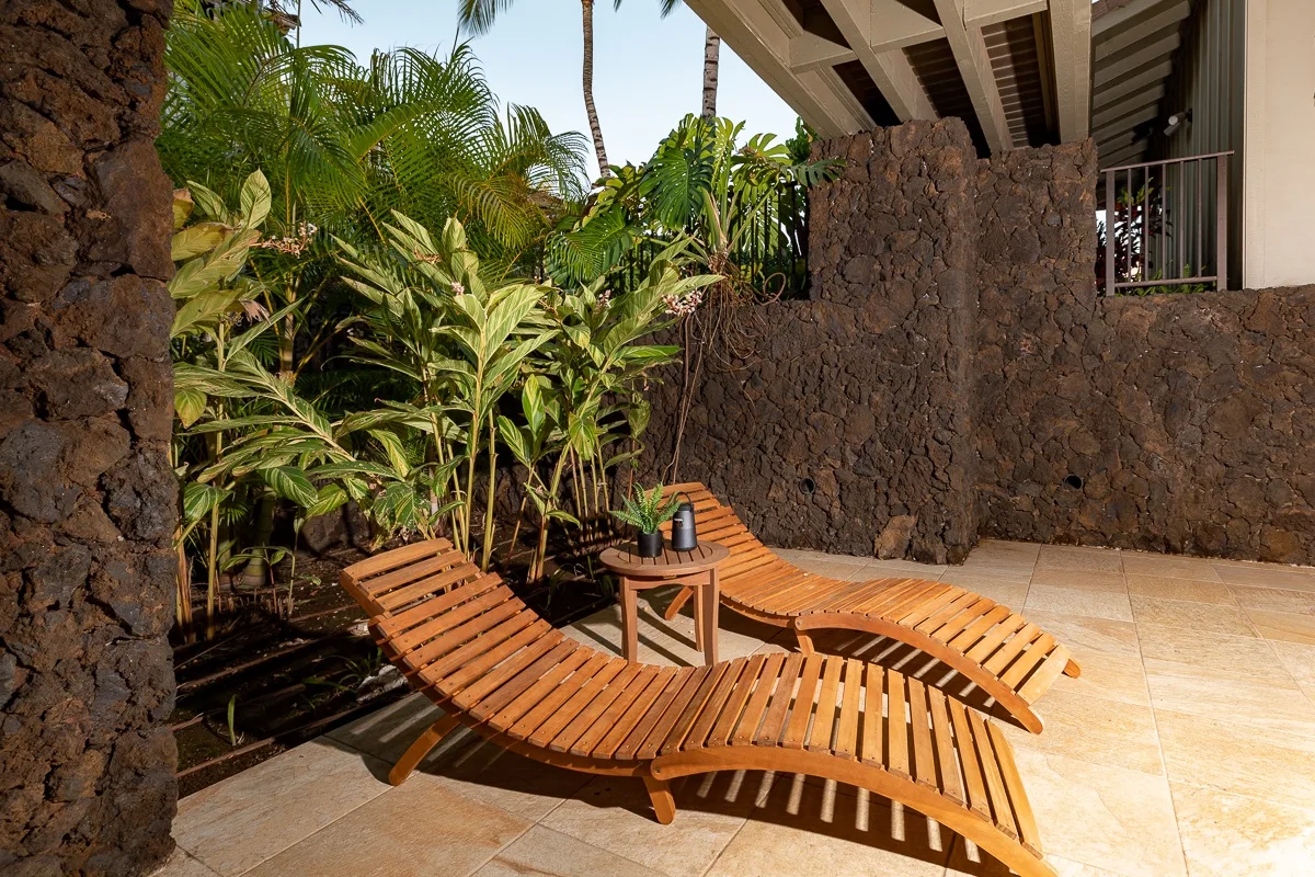 A sunny patio with wooden loungers beside tropical plants and a stone wall.