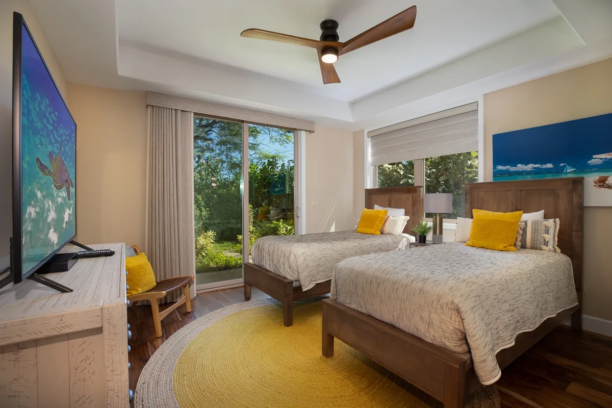 A bright bedroom with two beds, a television, and a view of greenery through glass doors.