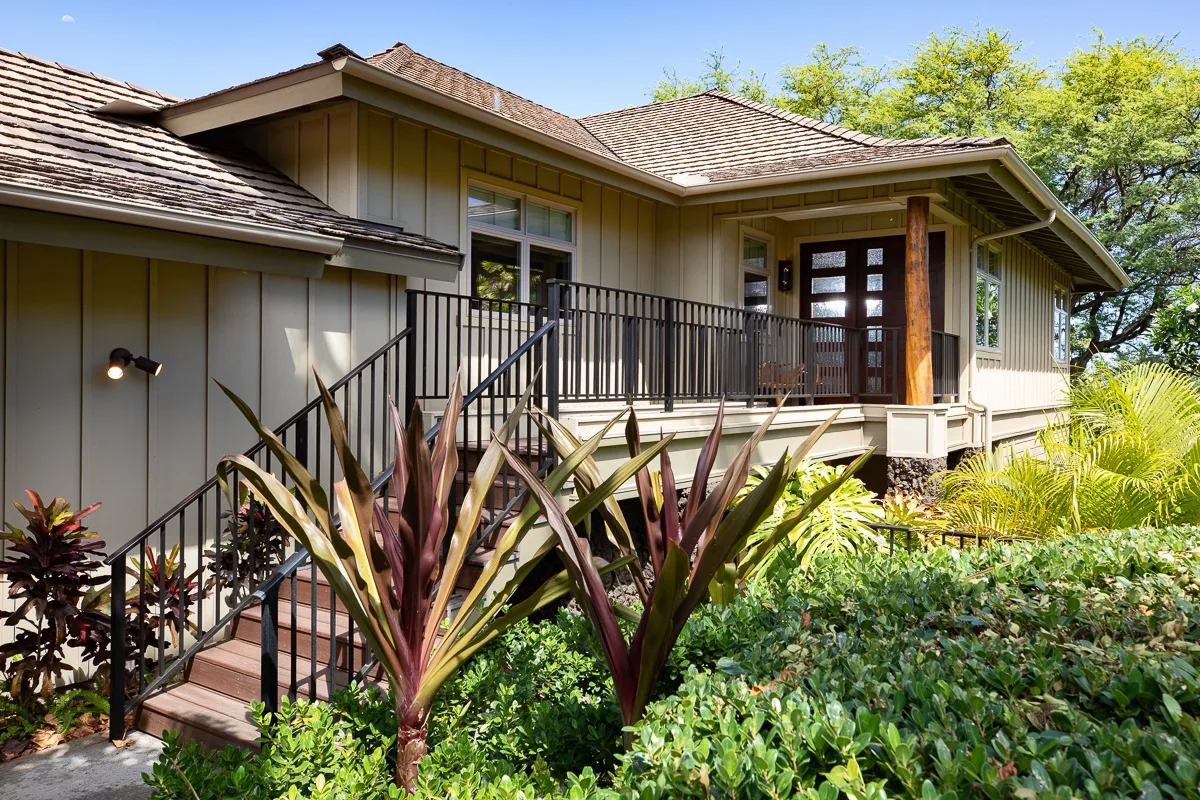 A house entrance with a raised wooden porch, dark railings, and surrounding garden plants.