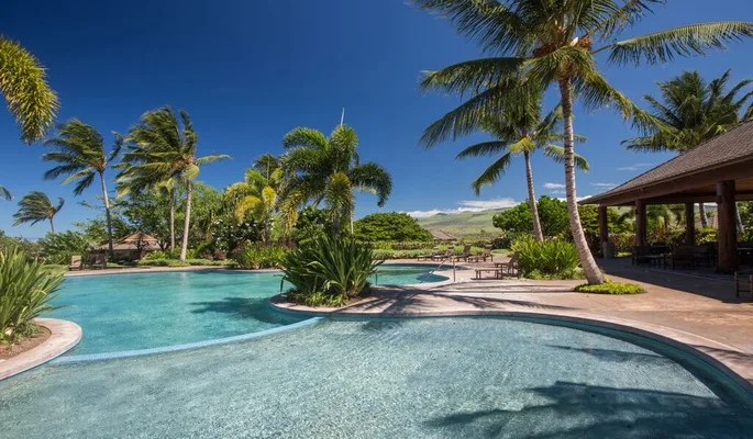 A tropical outdoor pool curves around a landscaped island beside a shaded pavilion.