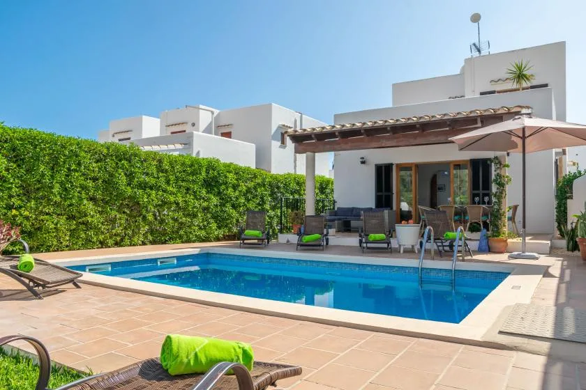 An outdoor swimming pool beside a modern white house, with patio seating and a hedge.