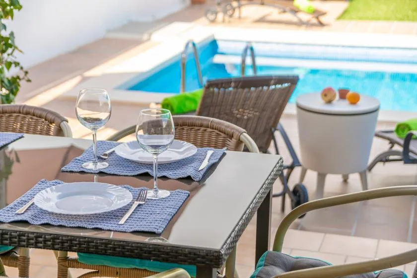 A poolside dining table is set with plates and glasses beside a swimming pool.