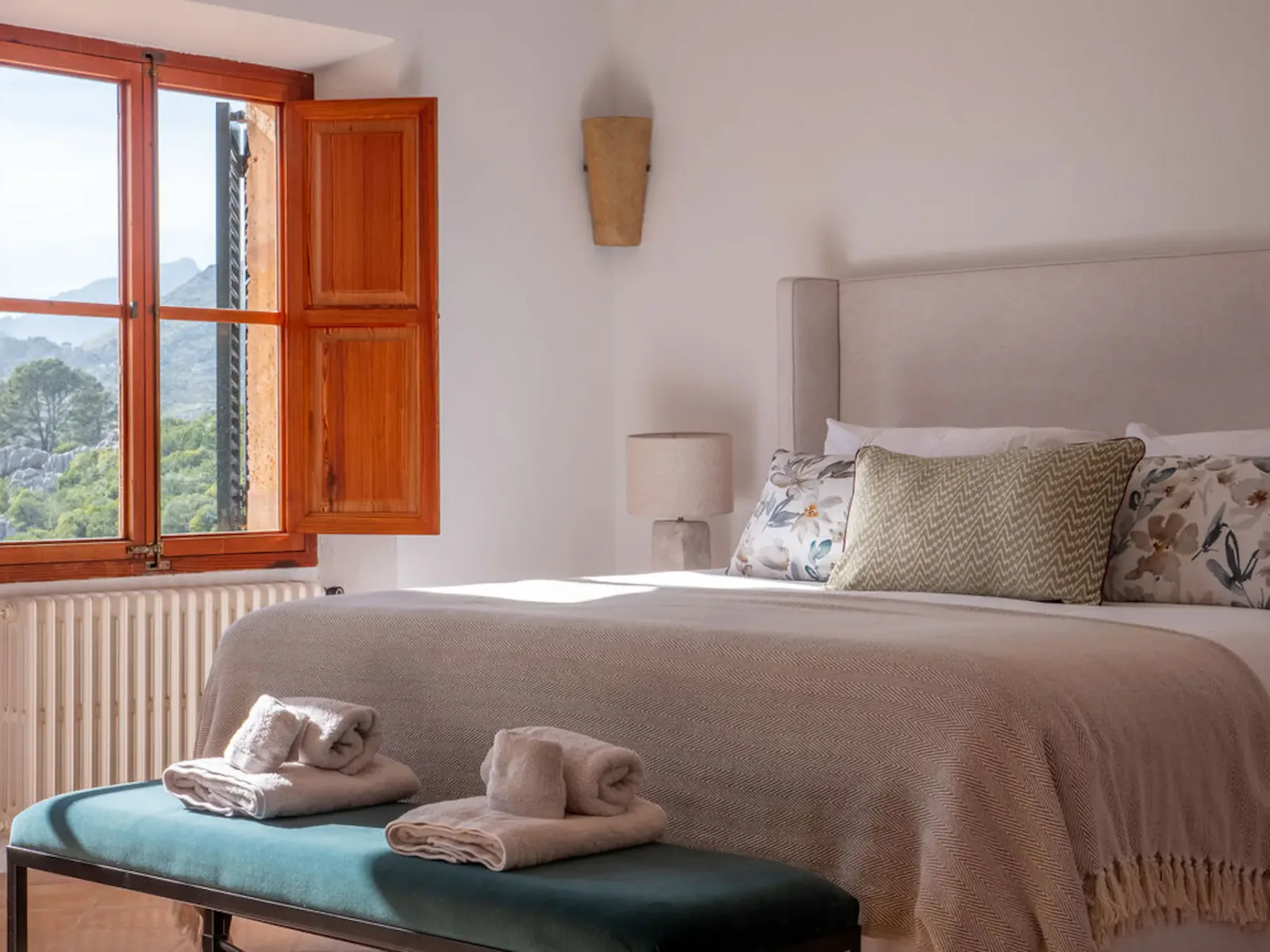 A bedroom with a neatly made bed and an open wooden window overlooking greenery.