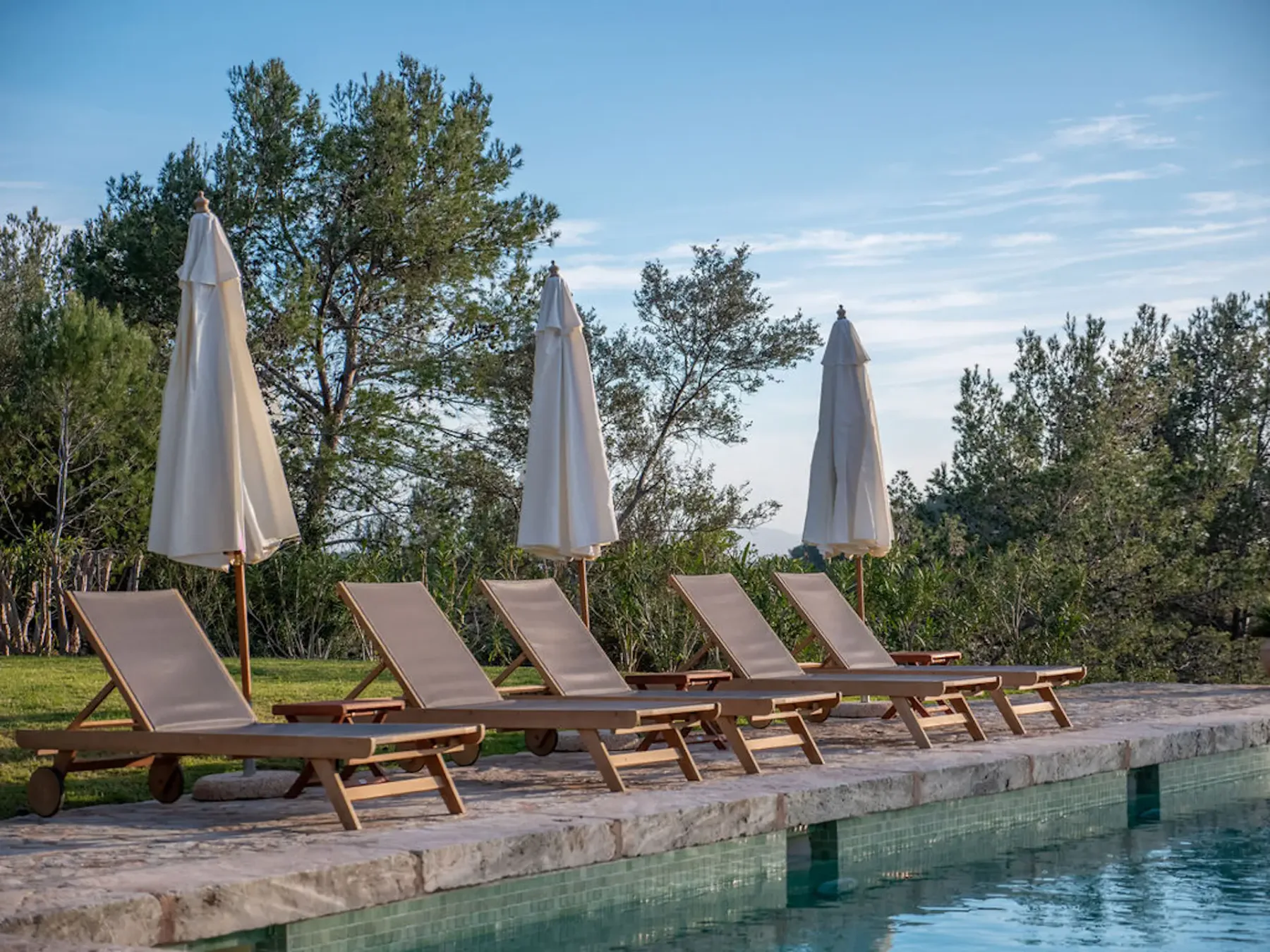 Poolside loungers sit beneath closed umbrellas beside a swimming pool.