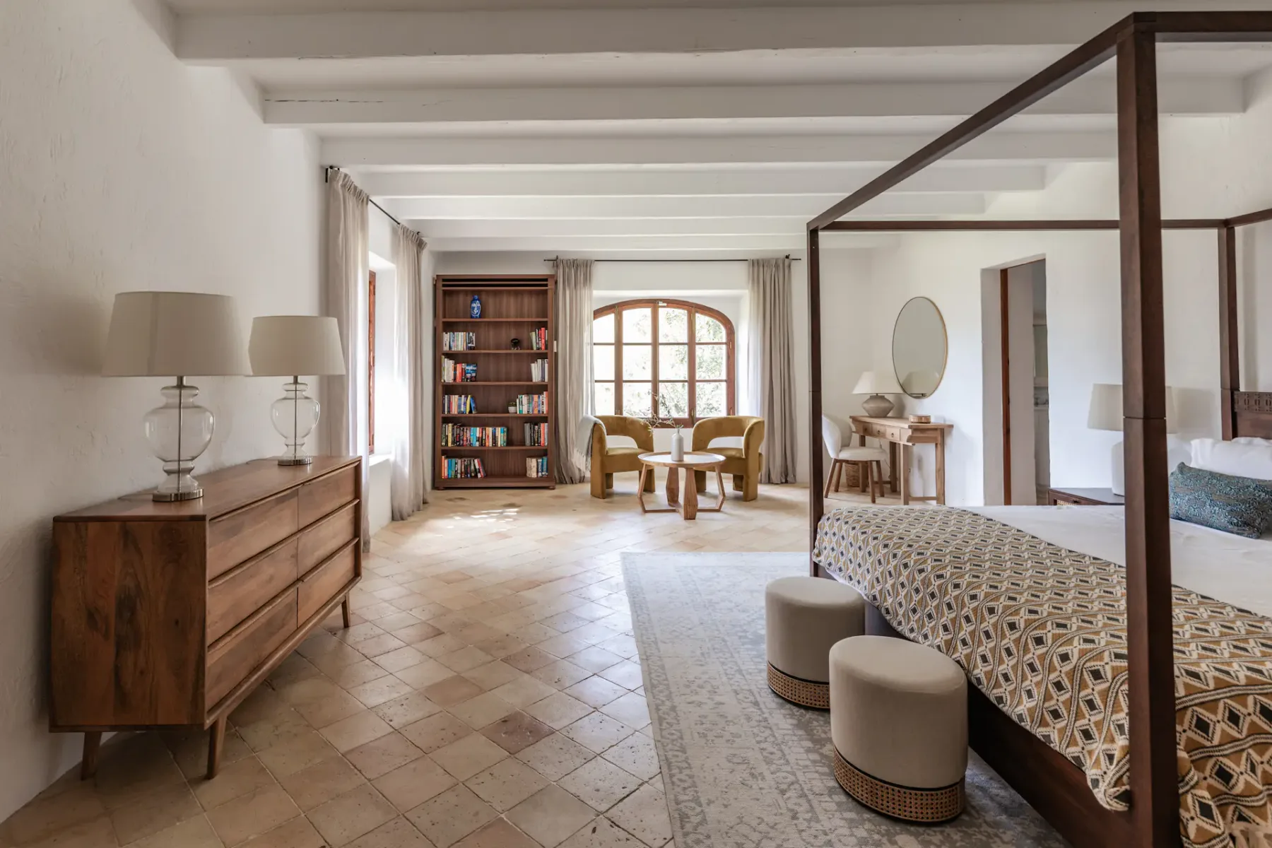 A bright bedroom with a canopy bed, wooden dresser, and seating by the window.