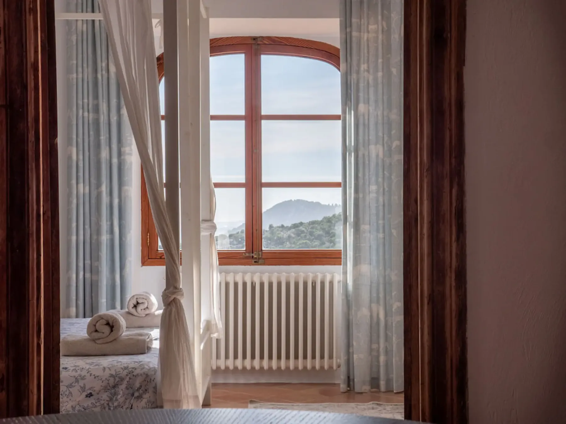 A bedroom with a bed beside a large window overlooking hills.