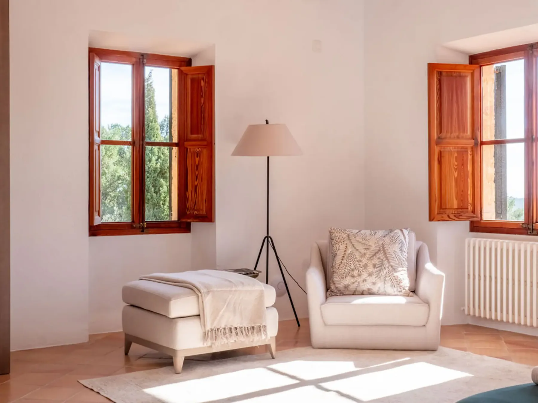 A bright sitting area with an armchair and ottoman beside open wooden windows.