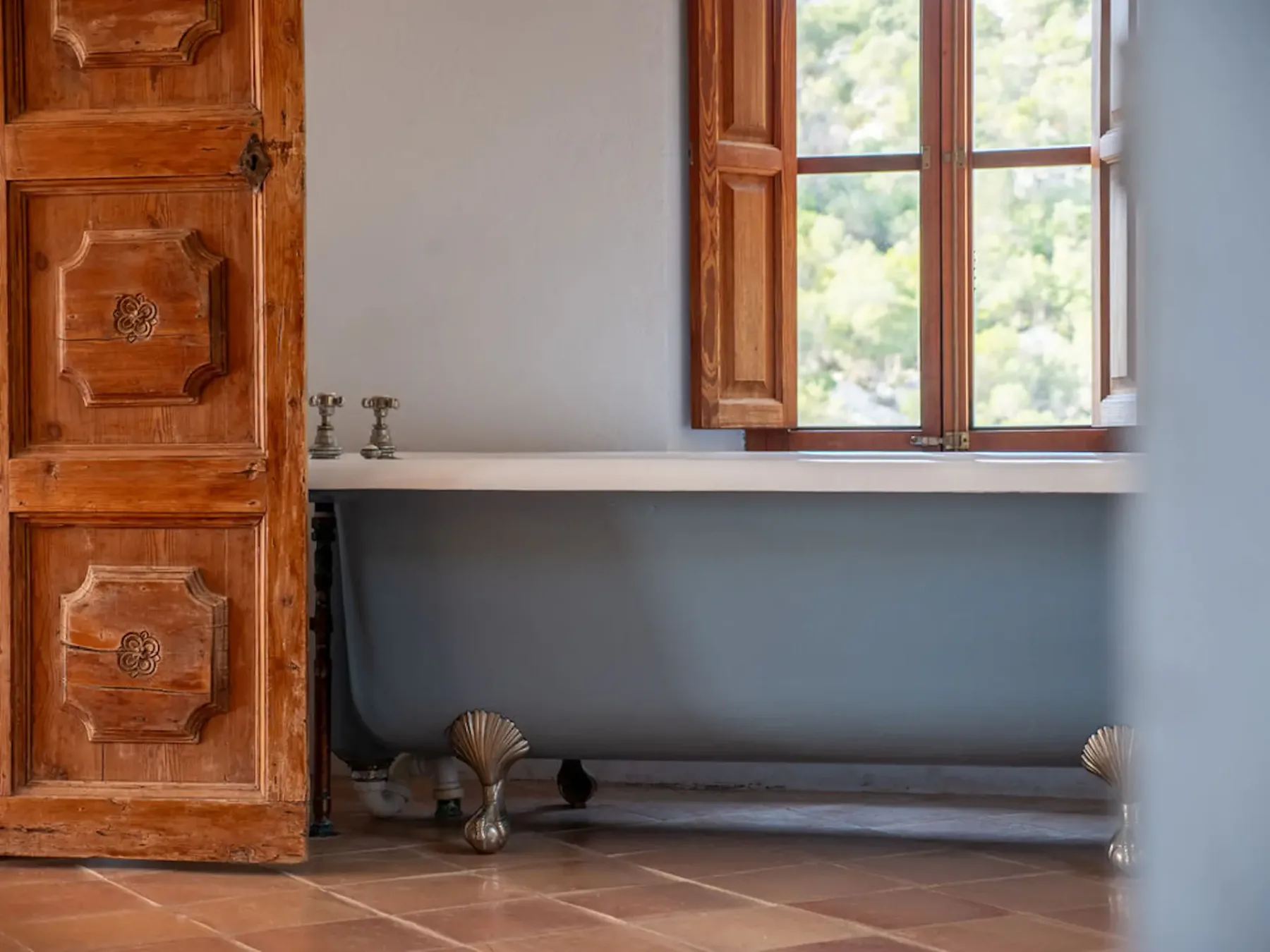 A bathtub sits beneath a wooden-framed window in a tiled bathroom.