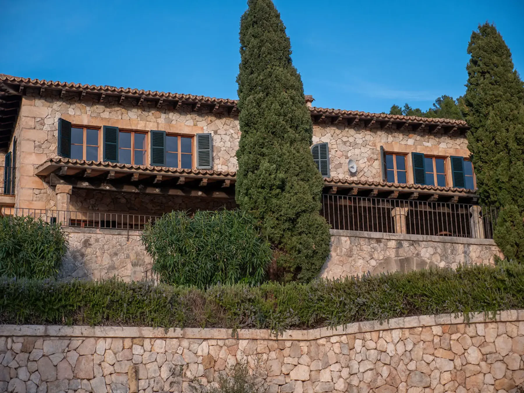 A stone house facade with shuttered windows and tall cypress trees.